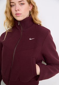 Burgundijos spalvos zip-up švarkas, pagamintas iš tekstūruotos medžiagos, turintis aukštą apykaklę, šonines kišenes ir baltą Nike logotipą priekyje.