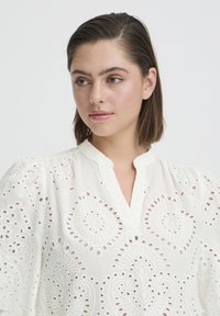 Femme aux cheveux bruns courts portant un chemisier blanc en œillet avec un col rond et des motifs découpés complexes, regardant légèrement sur le côté.