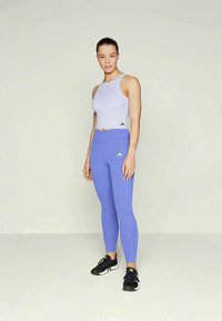 Nainen seisoo yksinkertaista taustaa vasten, päällä vaalea laventelinvärinen hihaton crop-top, siniset legginssit ja mustat urheilukengät.