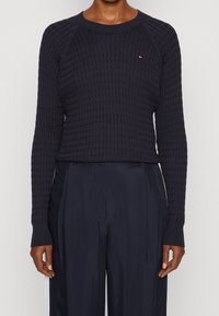 Dunkelblauer gestrickter Pullover mit strukturiertem Muster, langen Raglanärmeln und gerippten Bündchen. Verfügt über ein kleines Emblem auf der Brust.
