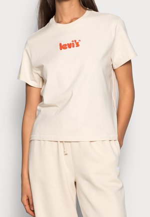 Beige bomulds t-shirt med korte ærmer, rund halsudskæring og rødt broderet "Levi's" logo på brystet. Sammen med beige træningsbukser.
