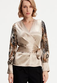 Haut enveloppant avec un corps en satin crème, manches longues à motifs noir et or, et un détail de nœud latéral ; texture lisse et coupe structurée.