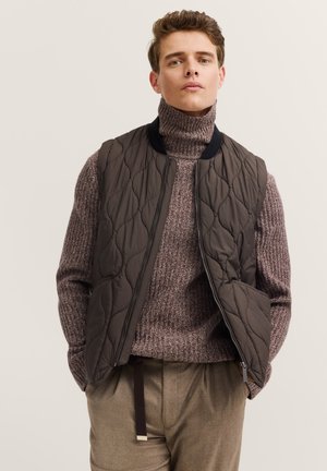 Gepolsterte braune Weste mit einem schwarzen Kragen, getragen über einem kräftigen beigen Rollkragenpullover. Sie verfügt über einen Reißverschluss und seitliche Taschen.