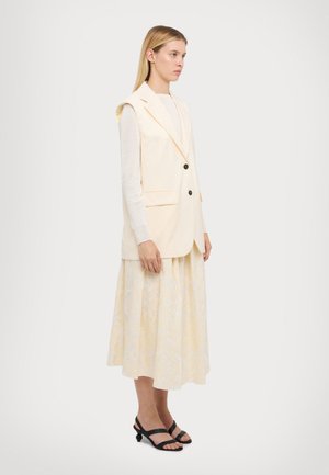 Gilet crema con revers e due bottoni neri, abbinato a una gonna a motivi gialla pallida e sandali neri con cinturini.