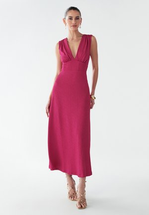 Fuchsia maxi dress with a deep V-neck and gathered shoulder details. Smooth texture, fitted bodice, flowing skirt, paired with strappy sandals.