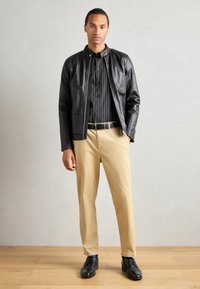 Black faux leather jacket over a black-striped shirt, paired with khaki pants and black shoes. Model stands against a neutral background.
