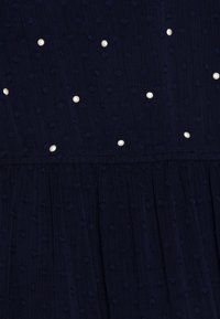 Marinblå klänning med texturerad tyg med upphöjda prickar och spridda vita pärlor, varsamt samlad vid sömmen för en extra detalj.