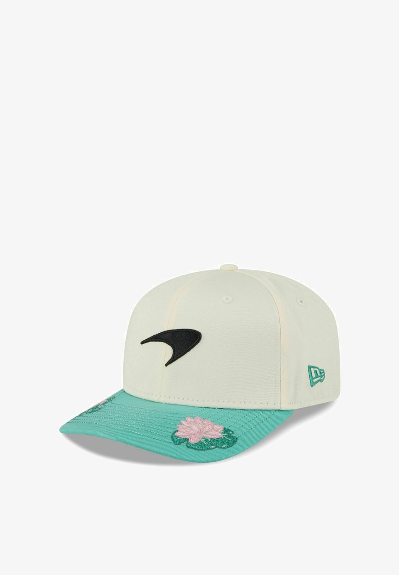 Casquette de baseball crème avec un logo noir, une visière turquoise ornée de broderies florales roses, et un tissu texturé. Elle comporte un détail avec une étiquette sur le côté.