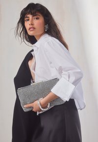 Vrouw met lang donker haar, gekleed in een wit shirt en een zwarte rok, houdt een clutchtasje met strass-steentjes vast, geassorteerd met een zilveren armband en een bloemvormige oorbel.