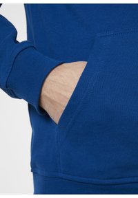Tejido de sudadera azul con una textura suave, que cuenta con un bolsillo frontal y puños acanalados. Primer plano de una mano descansando en el bolsillo.