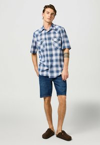 Lyhythihainen ruudullinen paita sinivalkoisena, jossa on kaksi rintataskua. Yhdistetty farkkushortsien ja ruskeiden puukiilakorkojen kanssa.