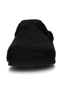 Scarpa nera senza lacci con una morbida texture pelosa, punta arrotondata e un dettaglio decorativo a nodo sul lato. Suola piatta e resistente per un comfort ottimale.