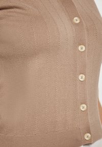 Ruskea urheilullinen neulecardigan, jossa on istuva malli. Edessä on pyöreitä vaalean beigejä nappeja. Tekstuuri näyttää pehmeältä ja joustavalta.