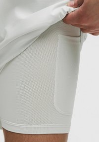 Pantalones cortos deportivos blancos confeccionados con una tela de malla texturizada y con un bolsillo lateral. El diseño incluye bordes suaves y una silueta ajustada.