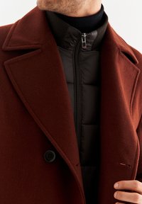 Strukturalny, brązowy wełniany płaszcz z szpiczastymi klapami, zapięciem na jeden guzik oraz czarną kurtką puchową założoną na spodzie, z widocznym zamkiem.