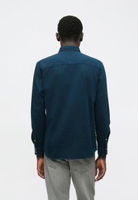 Man die met zijn rug naar de camera staat, in een donkerblauwe lange mouwen shirt en lichtgrijze jeans, tegen een eenvoudige witte achtergrond.