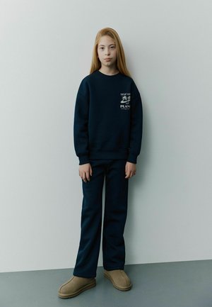 Granatowy sweter z logo na klatce piersiowej, w połączeniu z dopasowanymi spodniami. Model nosi beżowe klapki; gładki materiał, luźny krój i prosty fason nogawki.