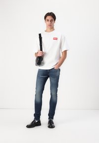 Fehér, rövid ujjú póló piros "DIESEL" logóval, elhalványult kék, slim-fit farmernadrággal és fekete bőr cipővel párosítva; egyszínű háttér.