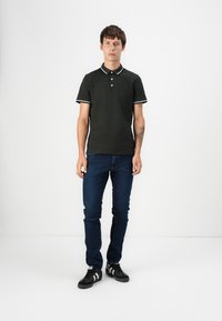 Polo-shirt vert foncé avec des accents blancs, deux boutons et des manches courtes, associé à un jean slim fit bleu foncé et des baskets noires.