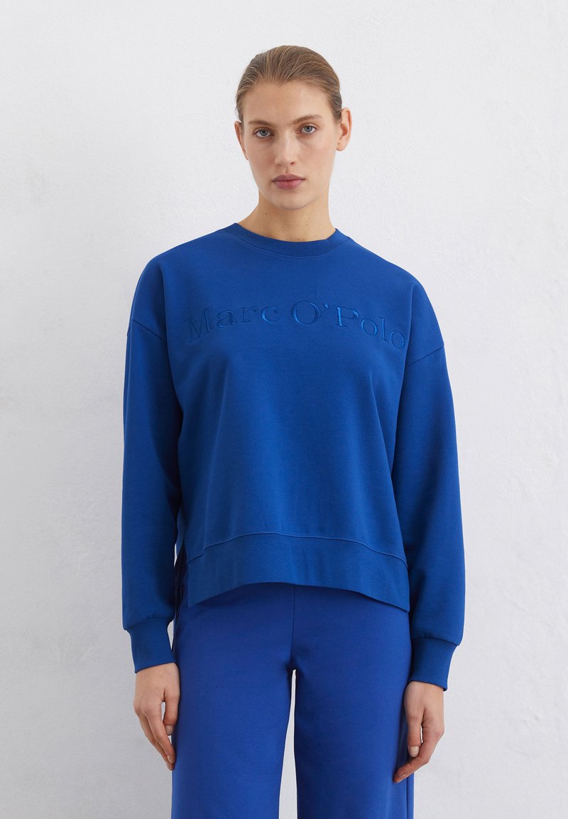 Blue sweatshirt with a crew neck, featuring embossed brand name "Marc O'Polo" on the front. Sweater has a relaxed fit and ribbed cuffs.