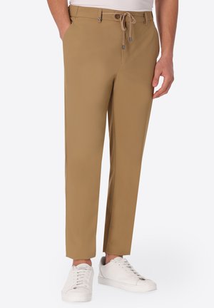 Uomo che indossa pantaloni slim fit color sabbia con coulisse in vita e sneakers bianche, in piedi con le mani parzialmente nelle tasche.