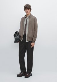 Brauner Strickcardigan über einem grünen Hemd mit Knöpfen, kombiniert mit dunkler Hose und braunen Lederschuhen. Hält eine schwarze Tragetasche.