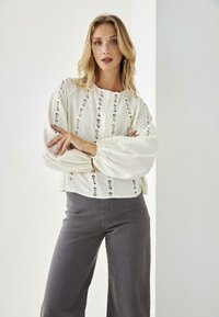 Blusa blanca texturizada con detalles de encaje y botones, combinada con pantalones de pierna ancha grises. La blusa tiene mangas abullonadas y un escote redondeado.