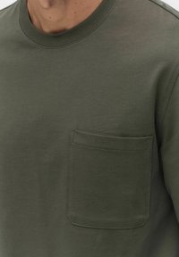 Olivgrön bomullströja med ribbad rund halsringning, med en bröstficka och slät yta. Inga synliga mönster eller metallaccenter.