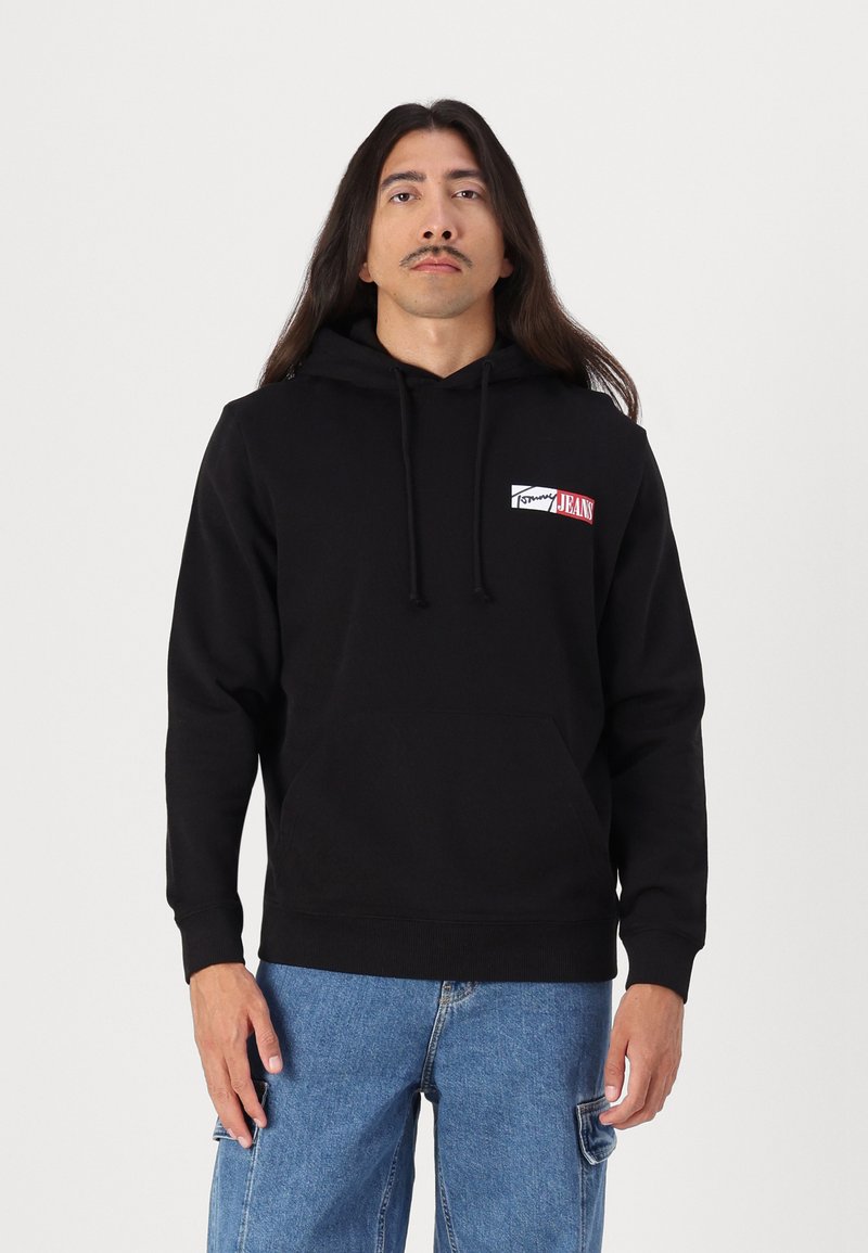 Sort hættetrøje med en frontlomme og snor, med et lille logo-patch i hvid og rød. Fremstillet af et blødt, struktureret stof.
