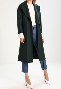 Lang, donkergroen jasje in een gestructureerd ontwerp met grote zakken, gedragen over een gebreide witte trui en blauwe jeans met zijstrepen.