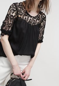 Sort bluse med blomstret blondeoverlay på ærmerne og halsudskæringen, lavet af letvægtsstof med A-linje form og pyntede detaljer.