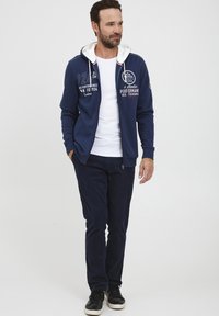 Tengerész navy zip-up kapucnis pulóver fehér béléssel, nyomtatott grafikával, sötét nadrággal és fekete sportcipővel párosítva. Mindennapi viseletre alkalmas, laza stílus.