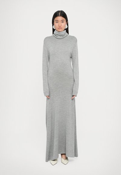 Vestido de punto gris con cuello alto, de manga larga, corte hasta el suelo y textura suave. Combinado con zapatos blancos de punta.