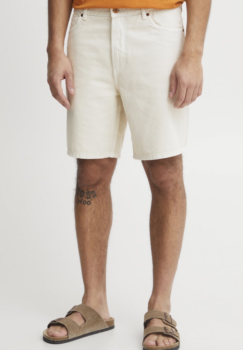 Shorts de mezclilla color crema con corte recto, bolsillos frontales y herrajes marrones. Llevados con sandalias marrones, que presentan una correa texturizada.