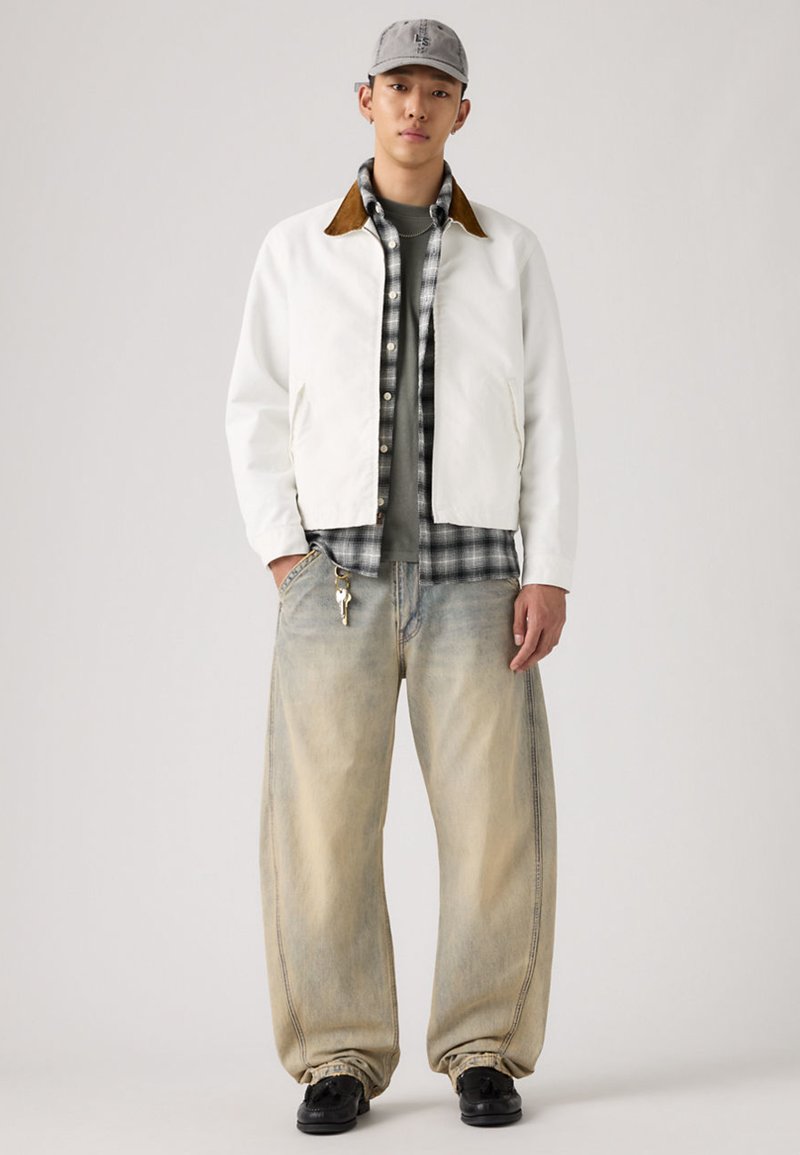 Giacca bianca con colletto marrone, camicia a quadri sotto, t-shirt grigia, jeans denim sbiaditi e larghi, e scarpe nere. Completa il look un berretto grigio.