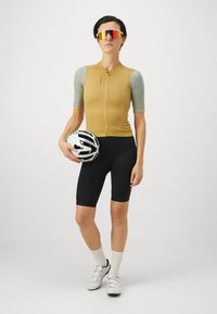 Fahrradausrüstung mit einem kurzärmligen, gold-braunen Oberteil und schwarzen Shorts. Zubehör umfasst einen weißen Helm und reflektierende Sonnenbrillen.