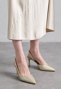 Talons slingbacks en paillettes dorées avec bouts pointus, un fin talon aiguille, et une finition en tissu lisse. Le design présente un dos ouvert.