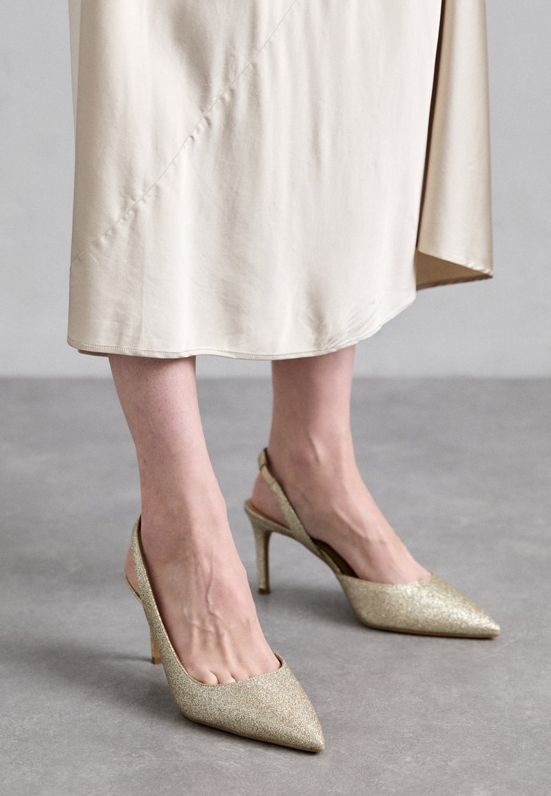 Talons slingbacks en paillettes dorées avec bouts pointus, un fin talon aiguille, et une finition en tissu lisse. Le design présente un dos ouvert.