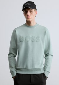 Világoszöld pulóver dombornyomott "BOSS" logóval, kerek nyakkivágással, bordázott mandzsettákkal és szegéllyel. Fekete sapkával viselve, amelyen fehér logóhímzés található.