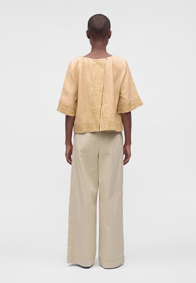 Personne vue de dos portant un haut beige avec des détails brodés et un pantalon large beige clair sur un fond uni.