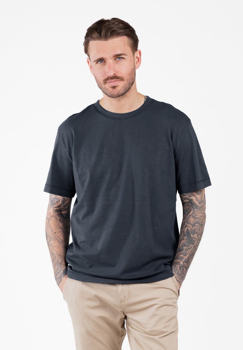 Man met tatoeages op beide armen, die een donkergrijs T-shirt en beige broek draagt, staand met handen in de zakken tegen een witte achtergrond.