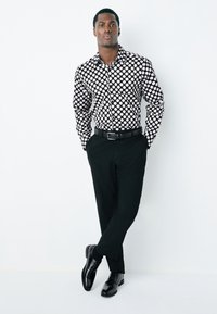 Camicia a pois bianca e nera con colletto, pantaloni neri e scarpe nere lucide, mostrati su uno sfondo bianco.