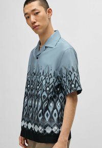 Camicia a maniche corte in tessuto blu chiaro con un motivo geometrico nero all'orlo. Presenta un colletto a camicia e bottoni neri.