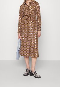 Brun mönstrad midi-klänning med långa ärmar, bälte i midjan, geometriskt tryck, bärs med silverfärgade loafers och en ljusblå handväska.