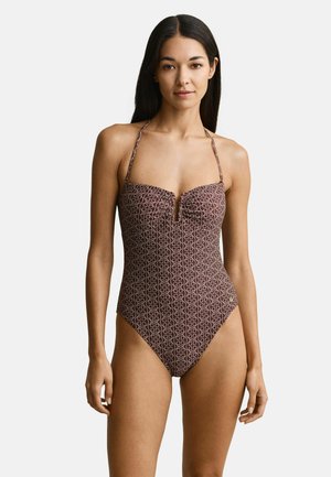 Kobieta w brązowym jednoczęściowym kostiumie kąpielowym z geometrycznym wzorem i paskiem na szyję, stojąca na tle jednolitego białego tła.