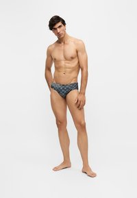 Jeune homme debout pieds nus portant un slip de bain sombre à motifs sur un fond clair uni.
