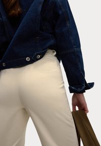 Jeansjacka i mörkblått med knappdetaljer, kombinerad med krämfärgade byxor. Hand som håller ett brunt föremål, som visar tygtexturer.