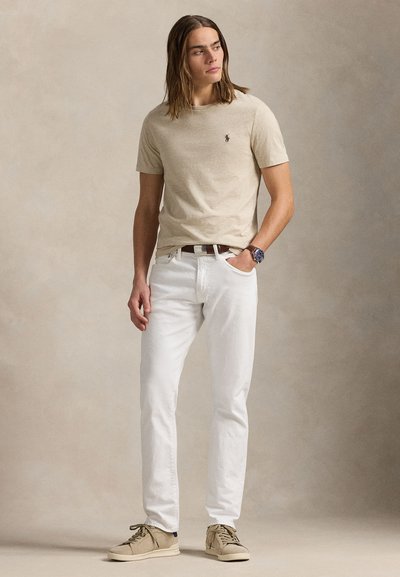 Mladý muž stojí, má na sebe béžovú košeľu s krátkym rukávom, biele rifle, tan tenisky, tmavý opasok a náramkové hodinky, na neutrálnom pozadí.