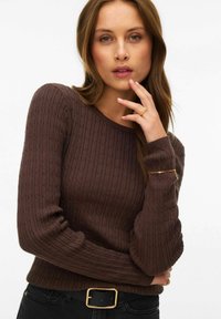 Femme aux longs cheveux bruns portant un pull en maille torsadée marron foncé et un pantalon noir avec une ceinture à boucle dorée, touchant son visage d'un air réfléchi.