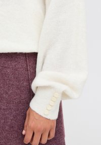 Pull en crème avec une finition douce et texturée. Doté d'un poignet roulé avec cinq boutons en crème, associé à une jupe en laine bourgogne foncé.